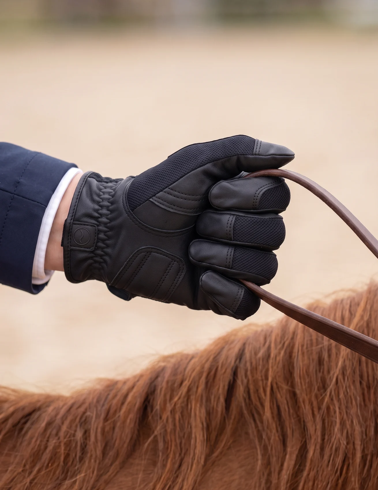 Mano izquierda de una amazona con guante de piel de cabra negro RE-AC-01 sujetando la rienda en la pista